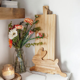 Wooden bunny-shaped cutting board and a rectangle-shaped cutting board with a vase of flowers and candle on a shelf.