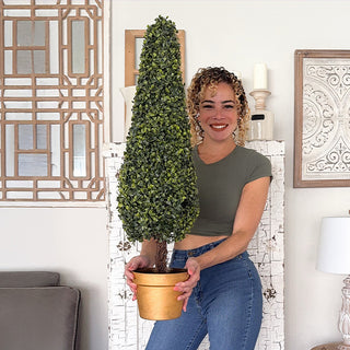 Woman holding tall faux boxwood topiary tree in gold pot inside modern home décor setting