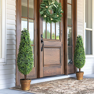 Pair of faux boxwood topiary trees in brown pots decorating a front porch with wooden door and wreath