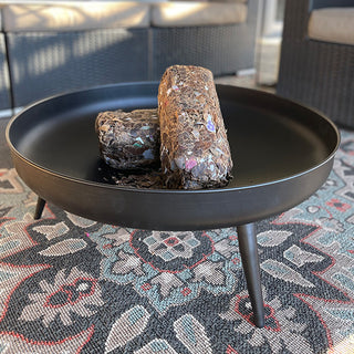 Black round metal fire pit bowl with two wood logs resting inside, placed on a decorative outdoor rug.
