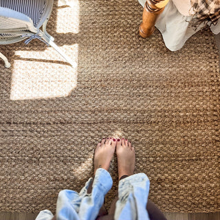 Feet on a brown, beige jute rug carpet with a chair and part of a person's leg visible.