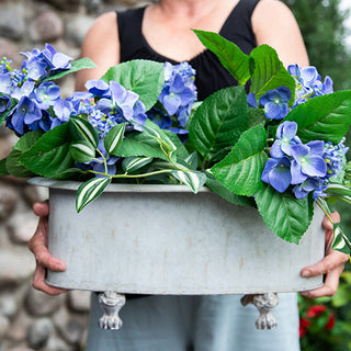 Set of two galvanized metal footed planters with blue hydrangeas displayed by stone fireplace
