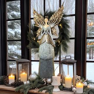 Holiday mantel display featuring a tall painted metal angel figurine with gold wings, surrounded by candles, pine garland, ornaments, and lanterns in a cozy winter home décor setting.