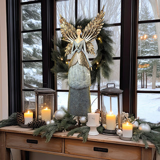 Christmas mantel styling with a tall painted metal angel sculpture accented by garland, candles, ornaments, and lanterns in front of large winter windows