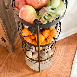 Overhead angle of a three-tier fruit storage rack showing apples, bananas, oranges, avocados, and potatoes organized in wire baskets.