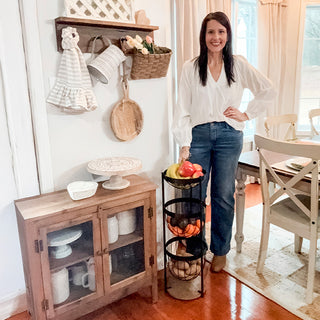 Woman in a farmhouse kitchen standing beside a fully stocked three-tier black metal fruit basket organizer.