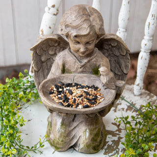 Angel statue bird feeder with birdseed in bowl surrounded by greenery