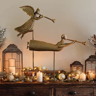 Rustic metal angel figurines with trumpets displayed on a wooden console table with candles, garland, and Christmas lights