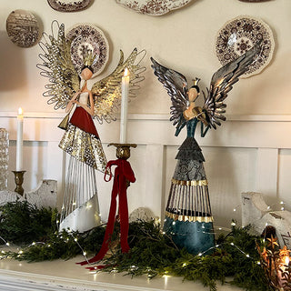 Two large handcrafted metal angel figurines displayed on white mantel with greenery and candles, featuring distressed gold and silver finishes