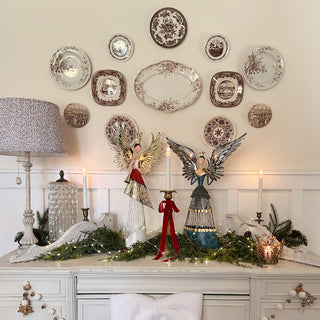 Christmas mantel decorated with metal angel figurines, transferware plates, white candlesticks, and evergreen garland