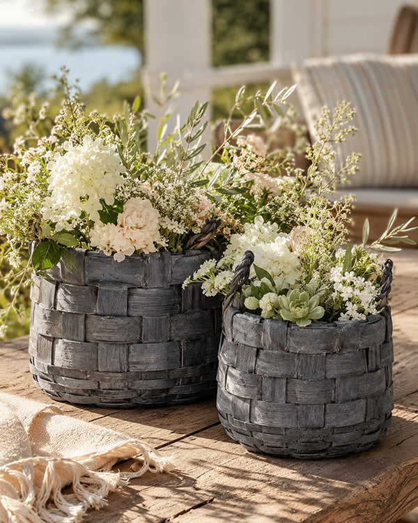Two woven stone planters with flowers on a wooden surface outdoors.