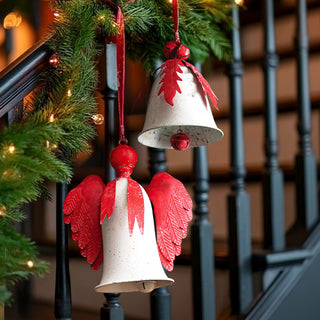 Rustic red and white distressed metal Christmas bell ornaments with holly leaves and angel wings hanging on staircase banister with evergreen garland