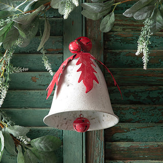 Close up of distressed metal holly and berry bell ornament with red painted leaf detail and jingle bell topper against rustic green wood backdrop