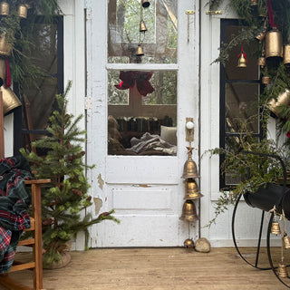 Rustic white door decorated with hanging brass bells, surrounded by greenery, holiday accents, and cozy porch styling