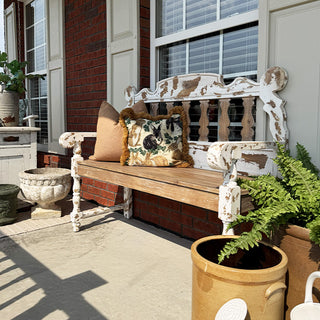 Wooden bench with decorative pillows on a porch, surrounded by potted plants.