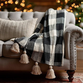 Black and cream buffalo check throw blanket with tassel detail draped over gray tufted sofa with cable knit pillow and Christmas lights bokeh background