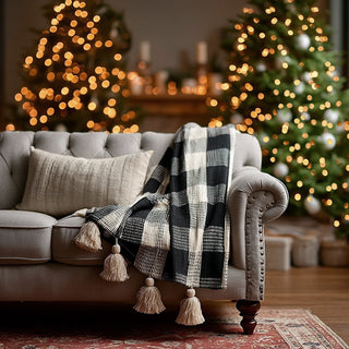 Farmhouse buffalo check throw with tassels styled on modern gray sofa in cozy living room with Christmas tree lights and neutral holiday decor