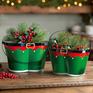 Set of two green metal elf buckets with red belt and gold buckle filled with pine branches and red berries on rustic wooden table with Christmas lights