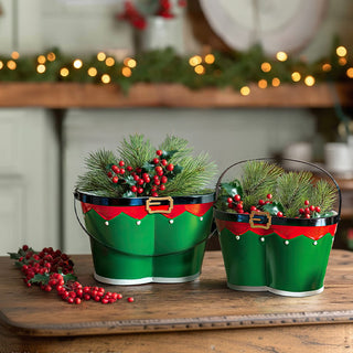 Christmas elf themed metal planters with Santa belt design holding holiday greenery and berries on farmhouse dining table with festive bokeh lights