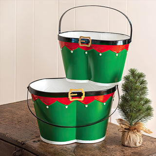 Two sizes of elf belt buckets displayed on wooden surface with small decorated Christmas tree showing metal handles for easy transport