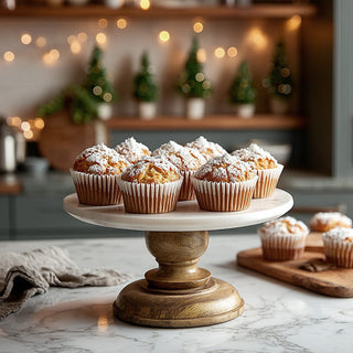 Decorative marble-top wooden cake stand holding assorted muffins in a cozy holiday-themed kitchen setting.