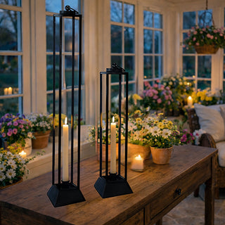 Set of 2 tall distressed black metal open frame pillar candle lanterns on rustic wooden table with flowers in sunroom