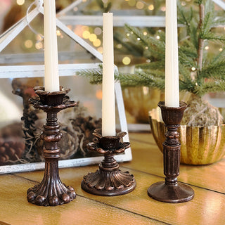 Three decorative candlesticks with candles on a wooden surface, with a blurred background of greenery and lights.