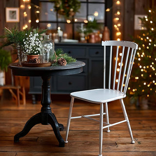 Distressed ivory metal farmhouse chair with spindle back next to black pedestal table in holiday decorated living room with Christmas lights