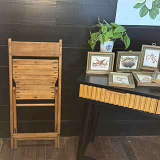 Folded vintage wooden chair leaning against a black wall beside a desk with framed art prints and a potted plant