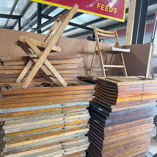 Group of stacked and displayed antique wooden folding chairs in various finishes inside a rustic warehouse setting