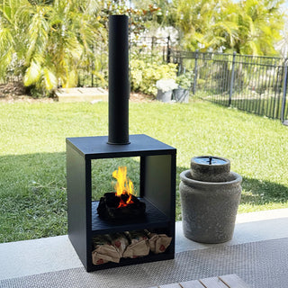Contemporary black chiminea with visible flames positioned on a backyard patio next to a stone water feature.
