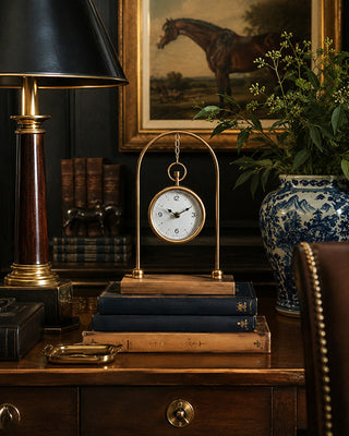 A brass and wood mantel clock with a round white face and black hands, displayed on a stack of books on a wooden desk in a classic study setting.