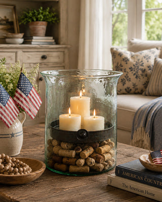 Decorative setup with candles in a glass container, American flags, and books on a wooden table.