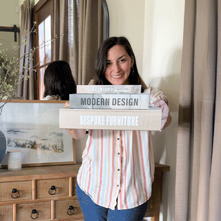 Woman holding a stacked set of decorative faux book storage boxes labeled Bespoke Furniture, Modern Design, and 19th Century Drawings in a styled home entryway