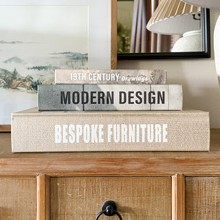 Close-up of stacked faux book storage boxes with titles Bespoke Furniture, Modern Design, and 19th Century Drawings used as decorative home accents.