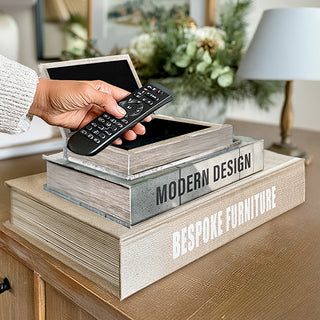 Hand placing a remote control inside a faux book storage box stacked with Modern Design and Bespoke Furniture titles on a home console table.