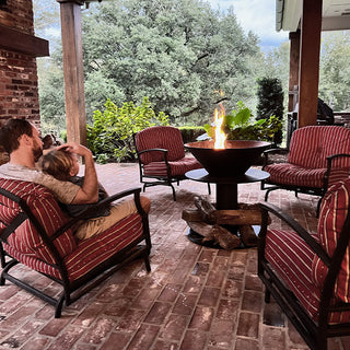 Covered patio with striped cushioned chairs arranged around a modern fire bowl as a man sits holding a child, creating a warm outdoor seating area
