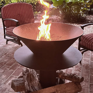 Modern black steel fire bowl with bright flame burning in the center of a brick patio, surrounded by red striped outdoor chairs