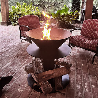 Iron fire bowl with stacked firewood and a tall flame, set on a brick patio with outdoor lounge chairs and lush garden landscaping.