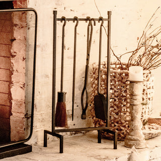 Black iron fireplace tool set displayed beside a rustic brick hearth, including broom, poker, tongs, and shovel, styled with cozy farmhouse decor.