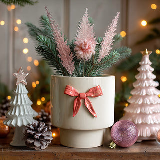 Festive ceramic Christmas planter with pink bow filled with sparkly winter greenery and pink flowers surrounded by white Christmas tree decorations and twinkling lights