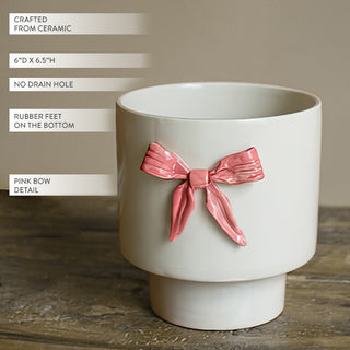 Cream ceramic planter with hand-painted dimensional pink bow detail shown on wooden surface highlighting 6 inch diameter and 6.5 inch height dimensions with no drain hole and rubber feet