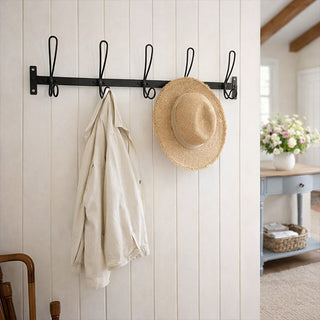 Black metal wall hook rack with 5 wire loop hooks mounted on white shiplap wall in farmhouse entryway with linen jacket and straw hat styled on hooks