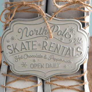 Close-up of embossed North Pole Skate Rentals metal sign reading ‘Hot Chocolate & Peppermints – Open Daily,’ tied with jute string on decorative skates