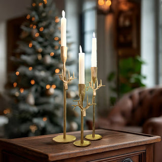 Set of three antique gold forged-iron taper candle holders on a wooden table with lit white candles, styled in a cozy holiday living room with a Christmas tree in the background