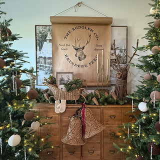 Holiday mantel decorated with Christmas greenery, wicker sleigh, rustic reindeer figure, and a set of antique gold taper candle holders, framed by Christmas trees and seasonal artwork