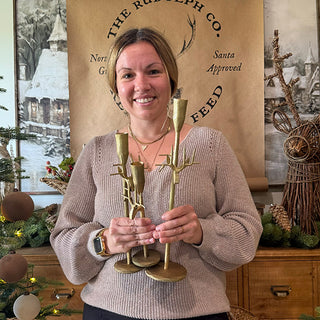 Woman holding a set of three gold reindeer-antler candle holders in a festive home decorated with rustic Christmas accents