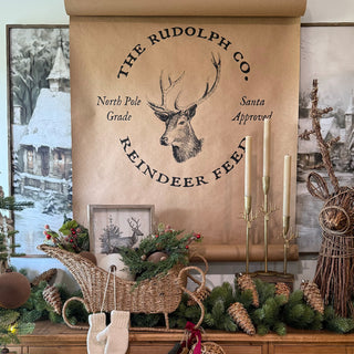 Rustic Christmas vignette featuring antique gold reindeer-antler candle holders displayed beneath a large Rudolph Co. reindeer feed wall scroll and surrounded by greenery