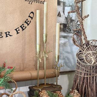 Set of three antique gold reindeer-antler candle holders with tall taper candles styled on a holiday mantel next to a wicker reindeer sculpture and winter artwork
