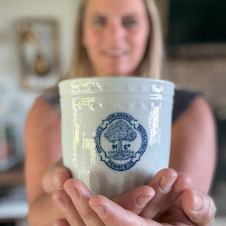 Woman holding vintage Belgian stoneware pot with blue “Confitures Mirland & Cie Frameries” logo, rustic kitchen decor background